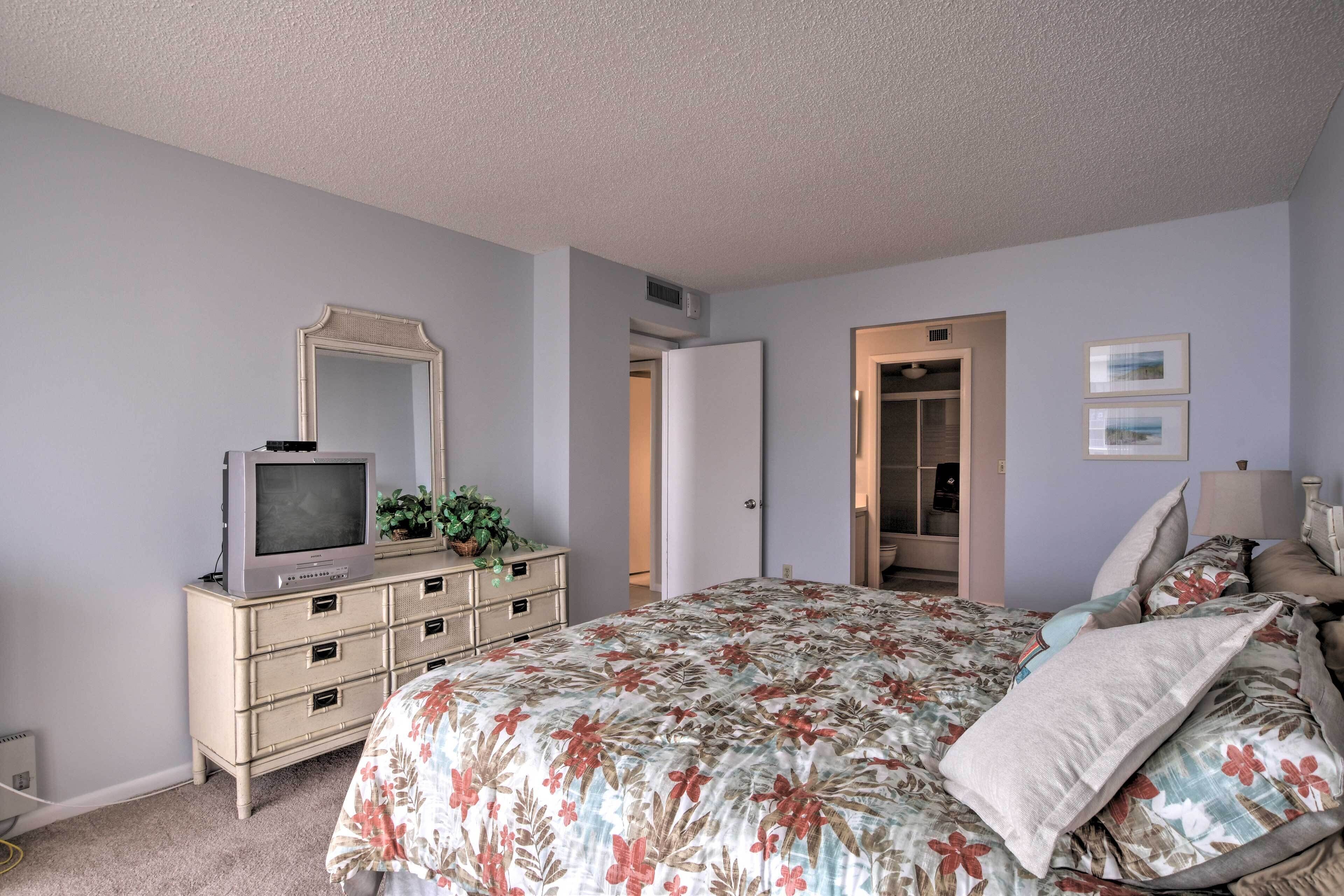 Une chambre à coucher comprenant un lit, une commode, un téléviseur et une porte donnant sur une autre pièce.