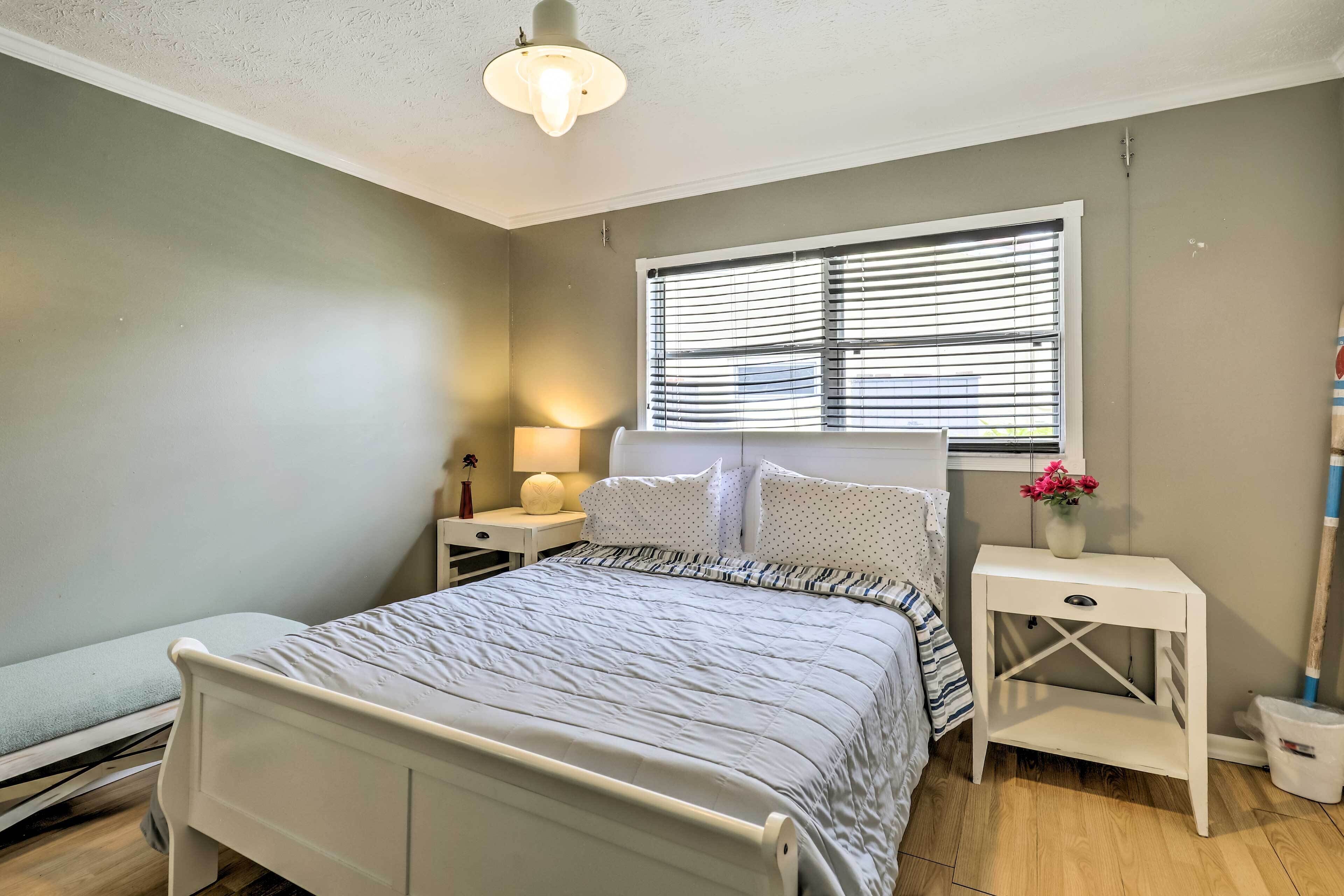 A bedroom with a bed, bedside tables, a window with blinds, a lamp, and a vase with flowers.