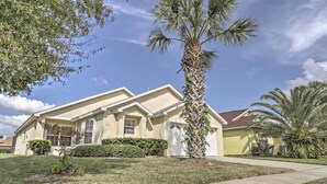 A two-story house with a beige exterior, a front yard with a palm tree, and a clear blue sky.