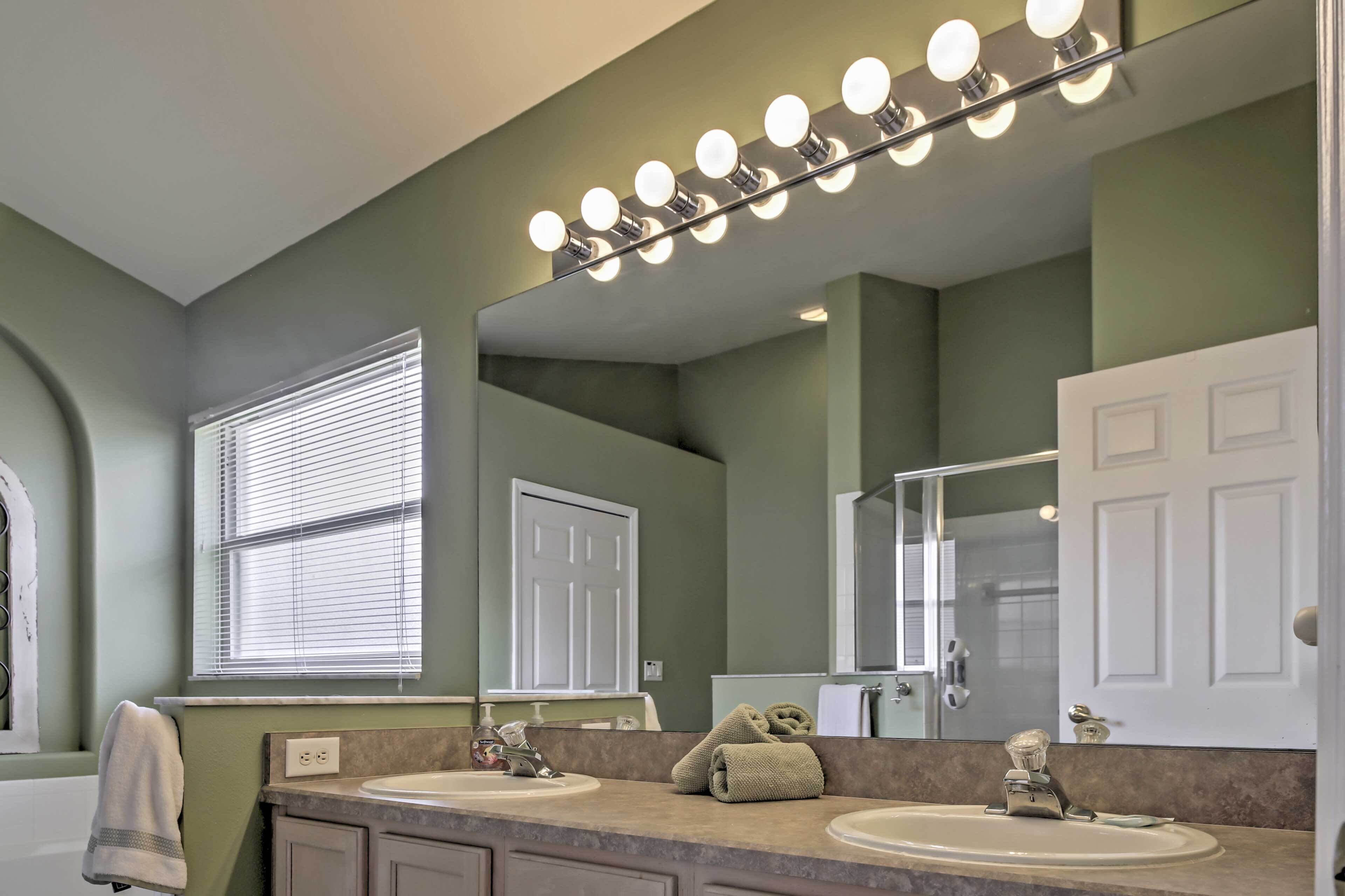 A bathroom with a vanity, two sinks, a mirror with lights, a towel rack, and a door.
