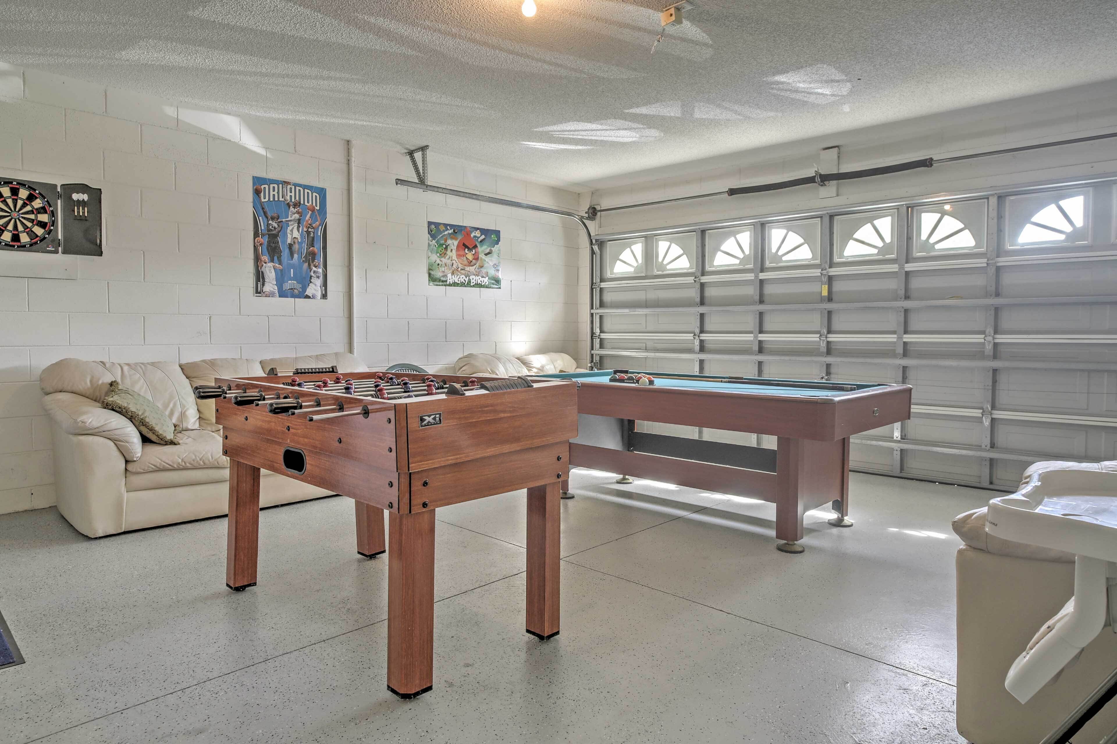 A room with a foosball table, a pool table, a dartboard, and a poster on the wall.