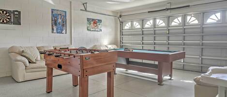 A room with a foosball table, a pool table, a dartboard, and a poster on the wall.