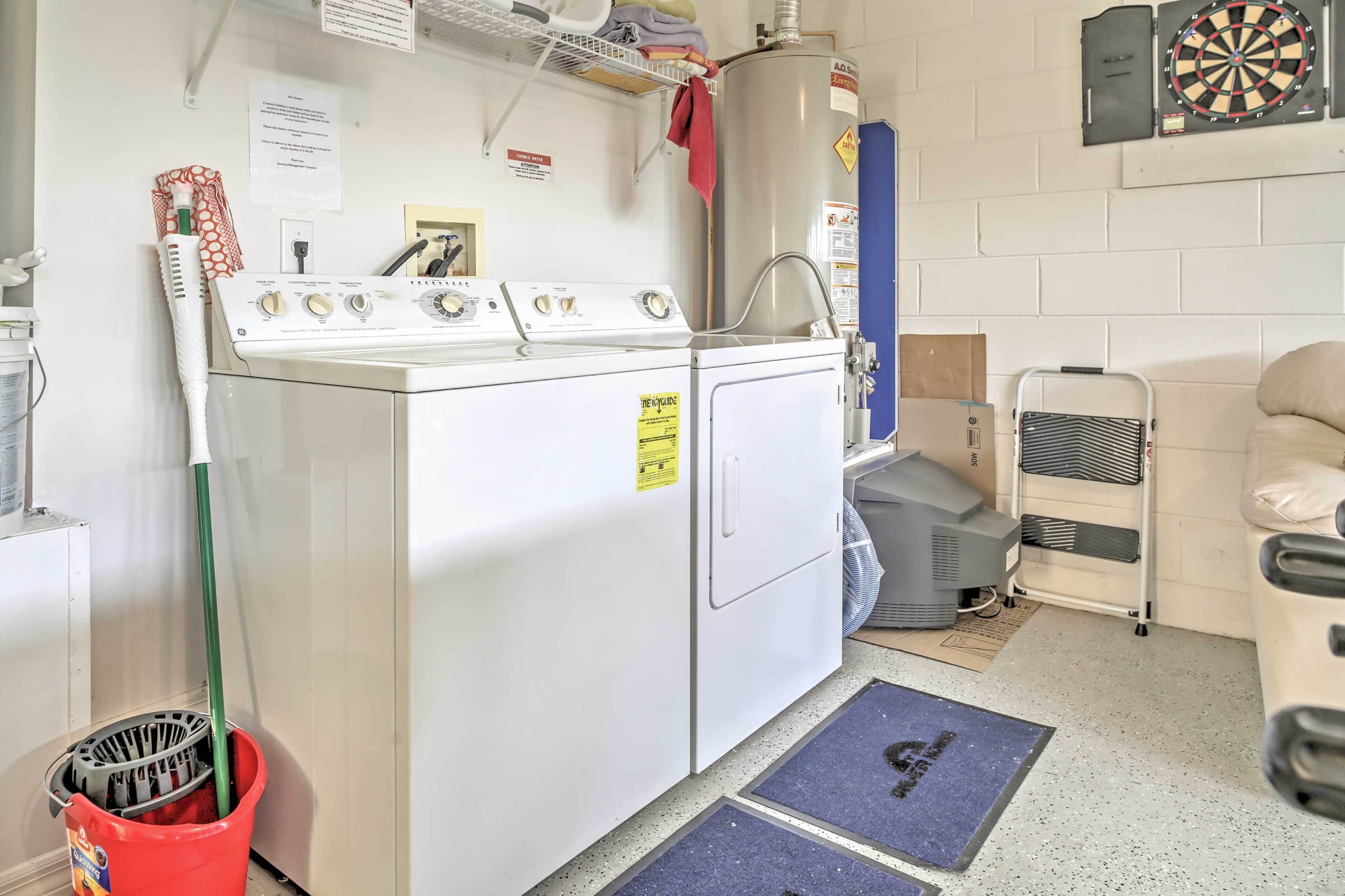 A laundry room with a washing machine, dryer, and ironing station. There is a mop bucket and a fan on the floor.