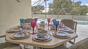 A covered outdoor dining area with a table set for a meal, surrounded by chairs, and overlooking a pool and trees.