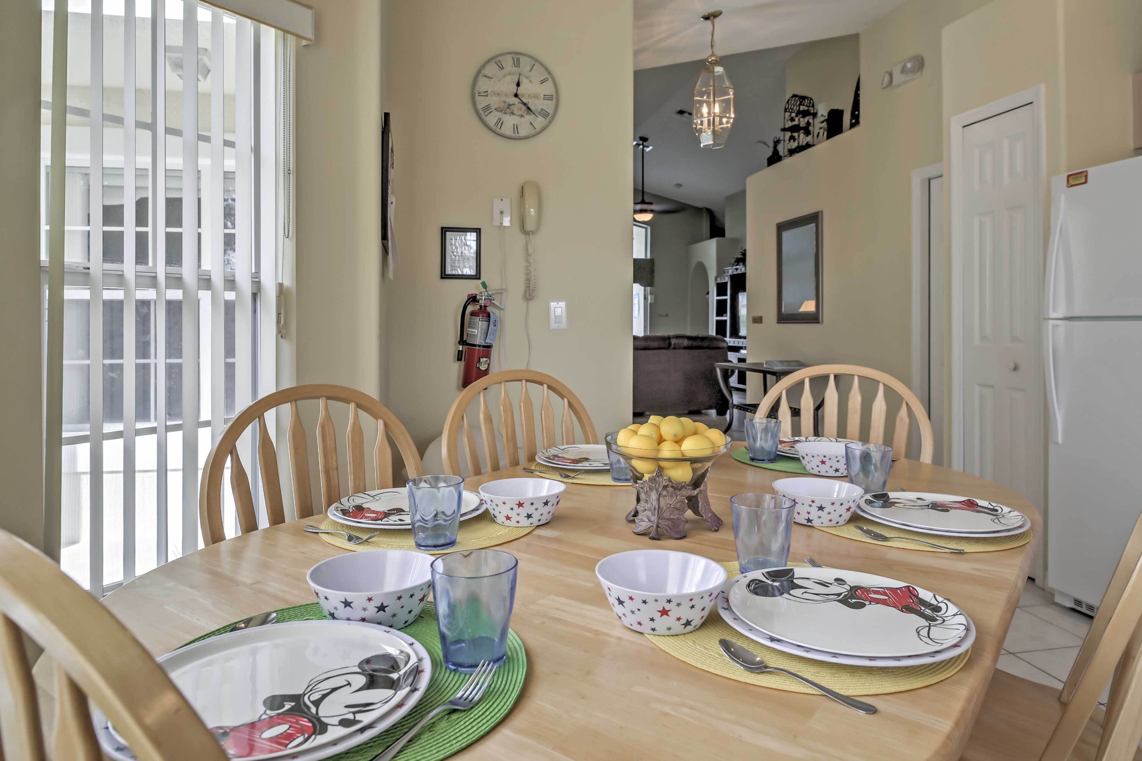A dining room with a round table set for six, surrounded by wooden chairs. A clock is mounted on the wall above the table.