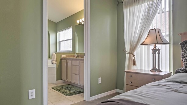 A bedroom with a bed, a nightstand, a lamp, a window with curtains, and a bathroom visible through an open door.