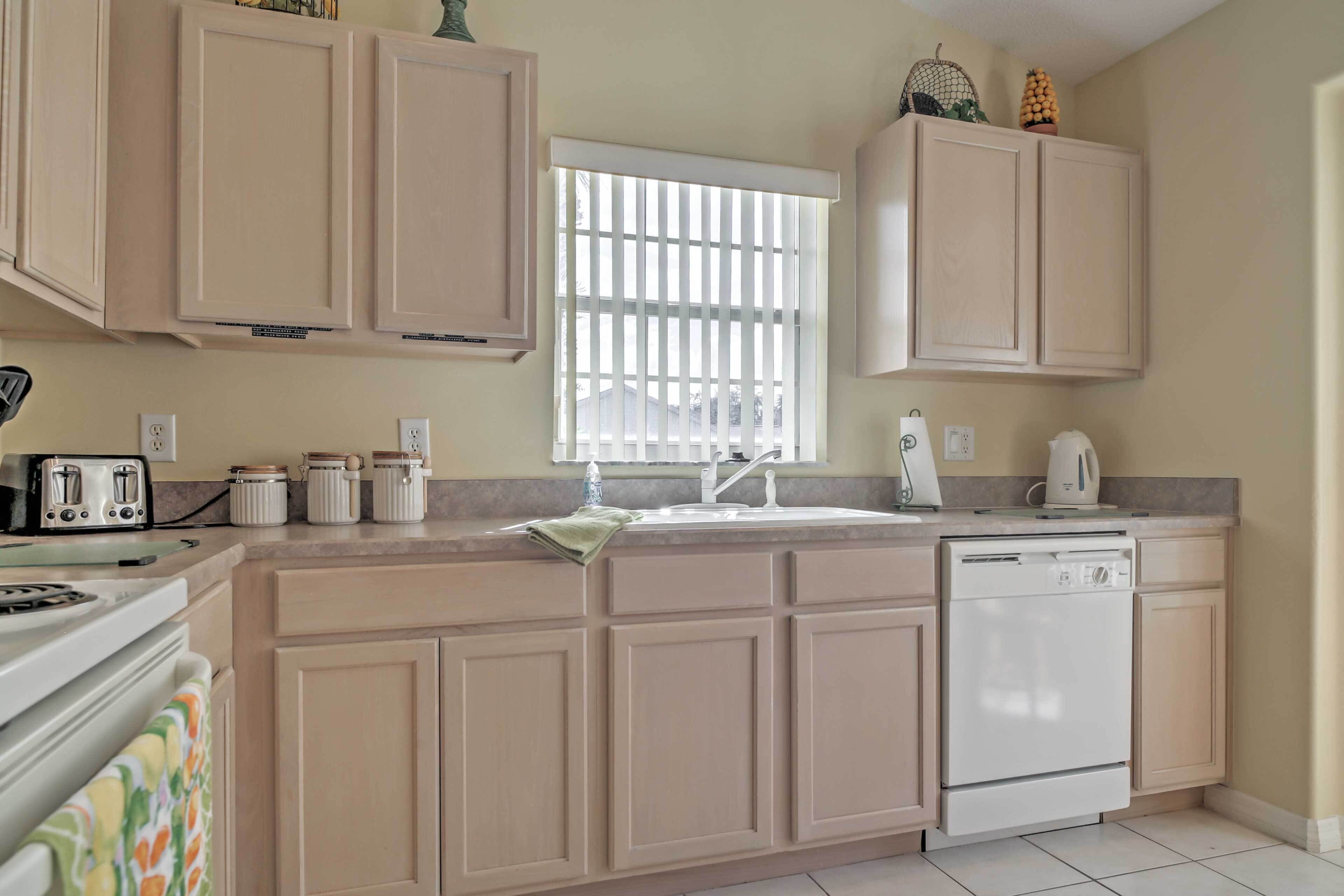 A kitchen with beige cabinets, a window with blinds, a stove, a dishwasher, and a countertop with various items.