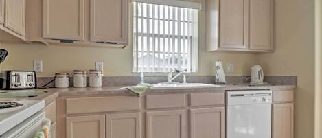 Une cuisine avec des meubles de couleur beige, une fenêtre avec des stores, une cuisinière, un lave-vaisselle et un plan de travail sur lequel se trouvent divers objets.