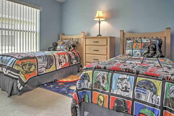 Une chambre avec deux lits, chacun recouvert d’une couette aux motifs de Star Wars. Il y a une table de chevet avec une lampe entre les deux lits, et une fenêtre avec des stores sur le côté gauche.