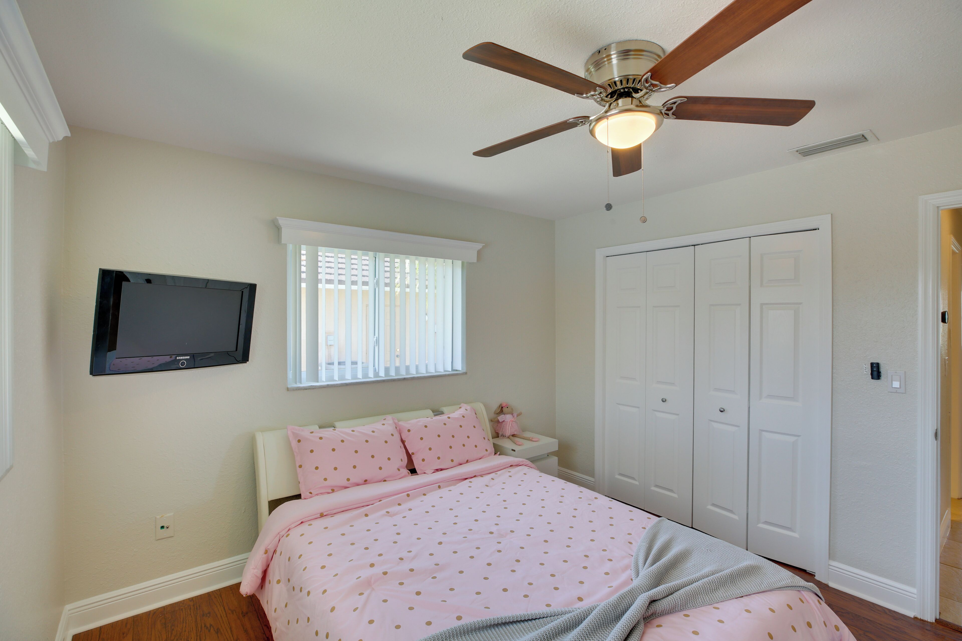A bedroom with a bed, a ceiling fan, a TV mounted on the wall, and a window with blinds.