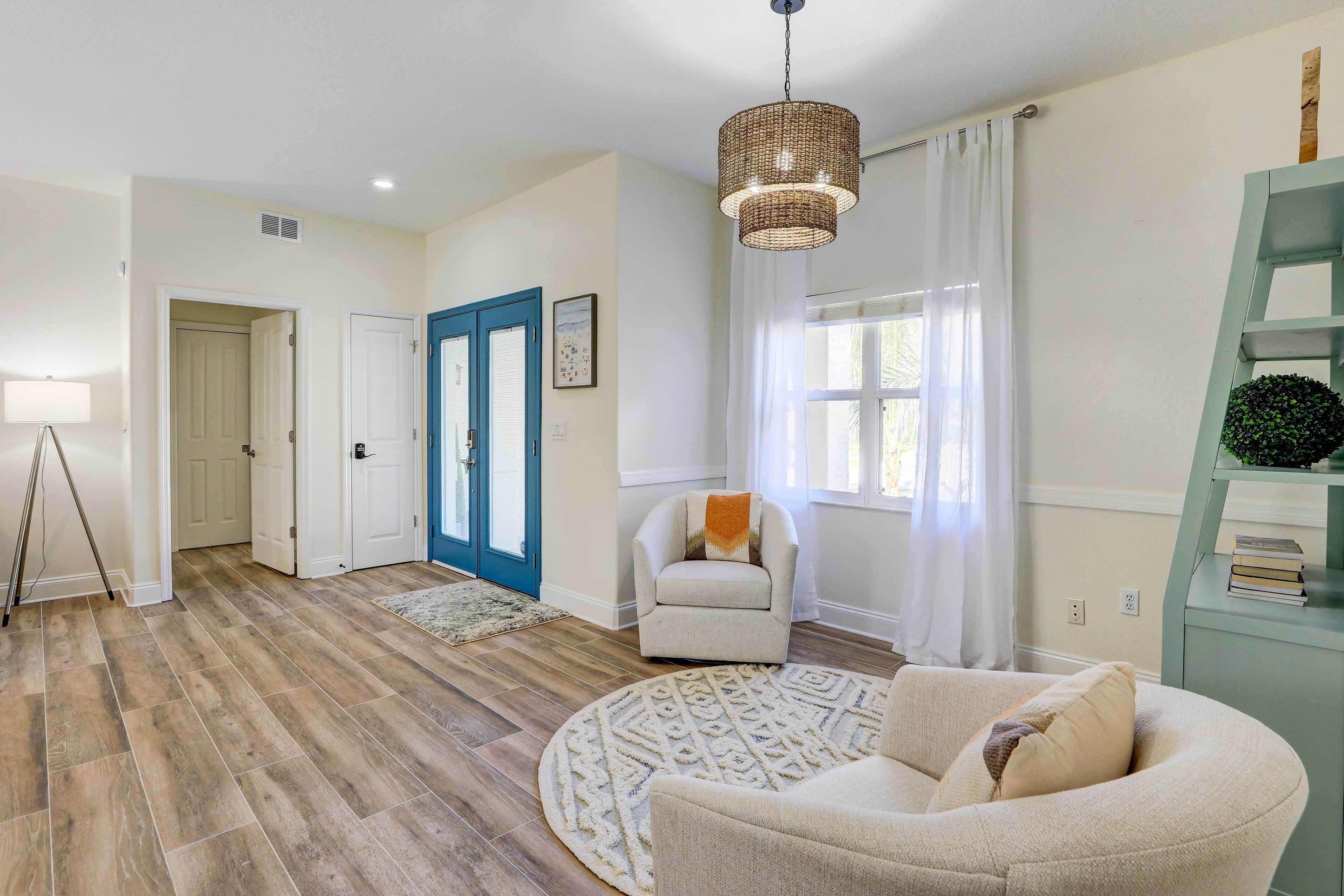 A spacious living room with hardwood flooring, a round rug, a beige armchair, a white armchair, a blue door, and a wicker pendant light.