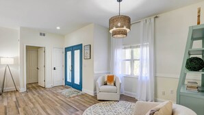 A spacious living room with hardwood flooring, a round rug, a beige armchair, a white armchair, a blue door, and a wicker pendant light.