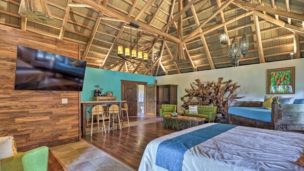 Ein geräumiges Zimmer mit einer hohen Holzbalkendecke, einem Bett mit blauer und weißer Bettdecke, einem grünen Sessel, einem Holztisch und einem an der Wand befestigten Fernseher.