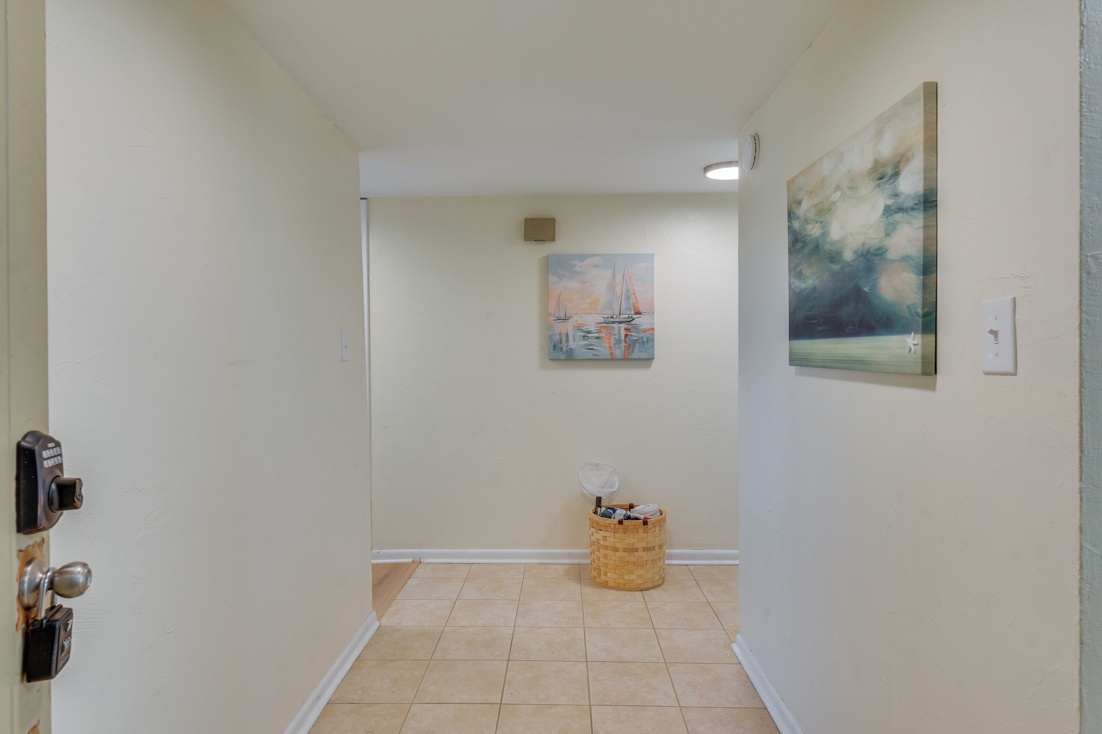 A hallway with a door on the left, a trash bin in the center, and two paintings on the walls.