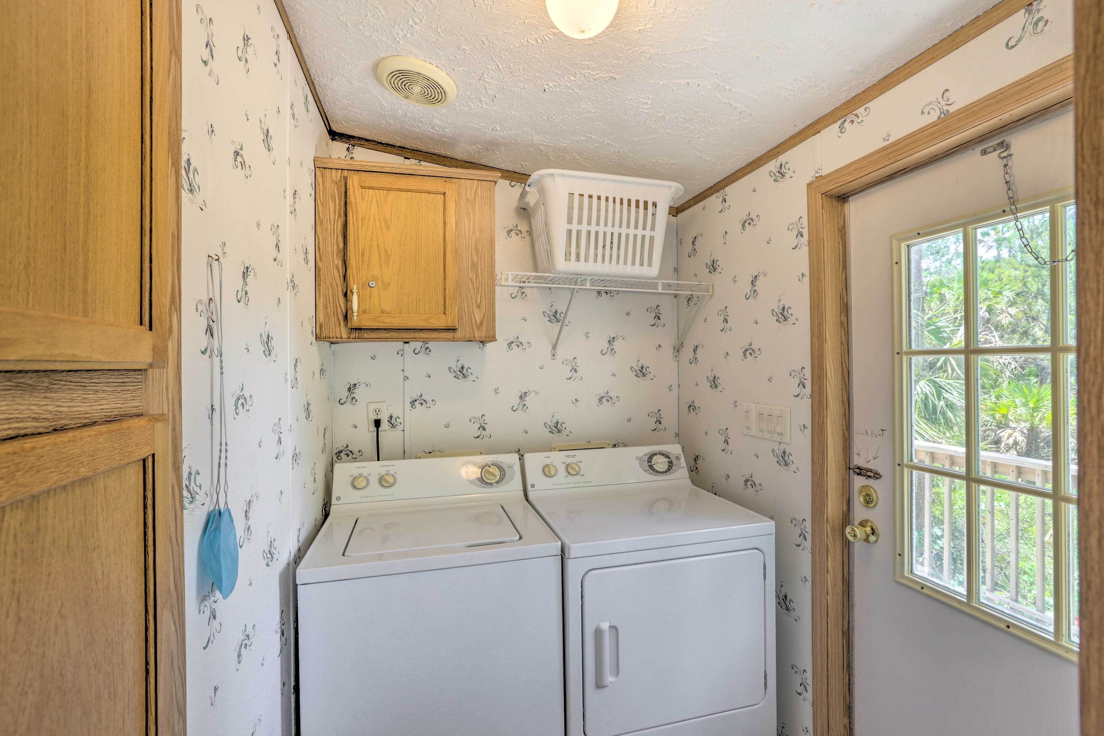 A laundry room with two white washers and dryers, wooden cabinets, a white laundry basket on a shelf, and a window with a view of greenery.