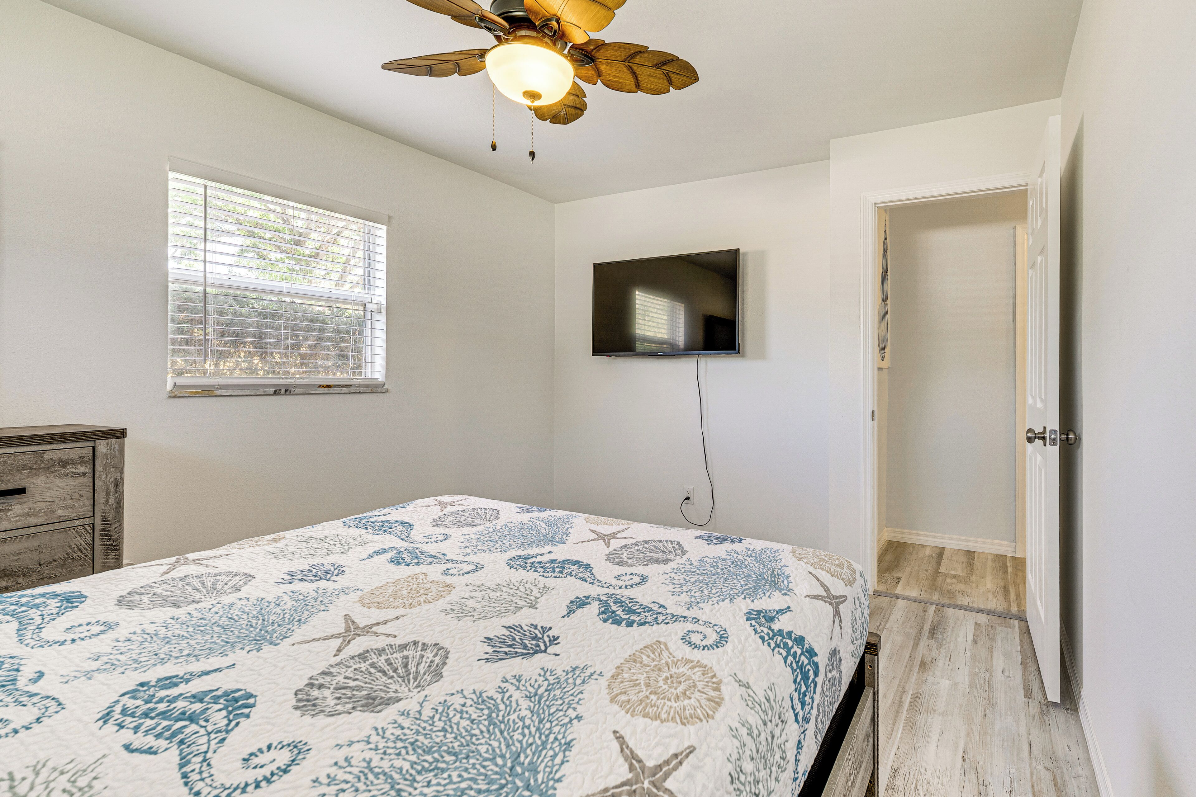 Une chambre à coucher avec un lit, un ventilateur de plafond, un téléviseur fixé au mur et une fenêtre avec des stores.