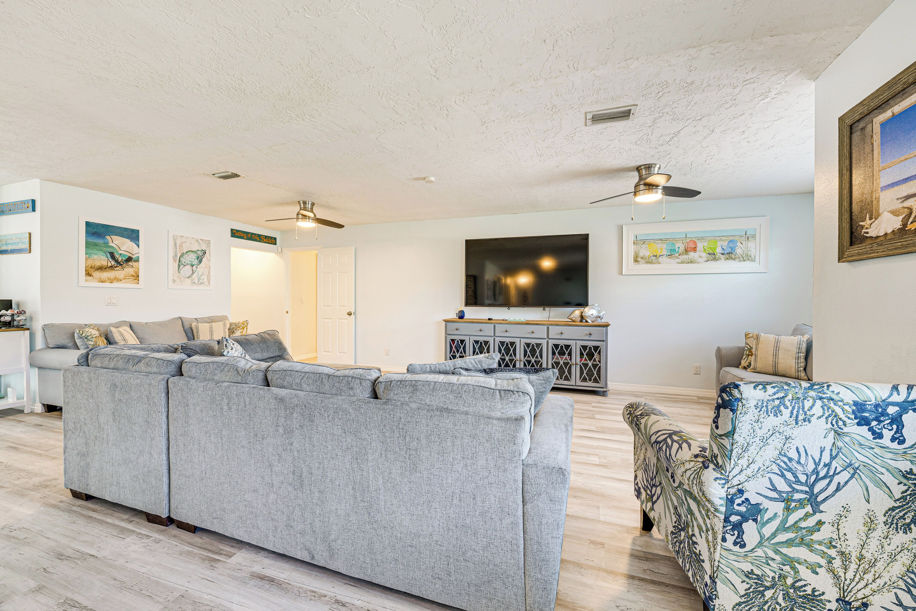 Ein Wohnzimmer mit grauem Ecksofa, einem gemusterten Sessel, einer Kommode mit Dekoration und einem an der Wand befestigten Flachbildfernseher.