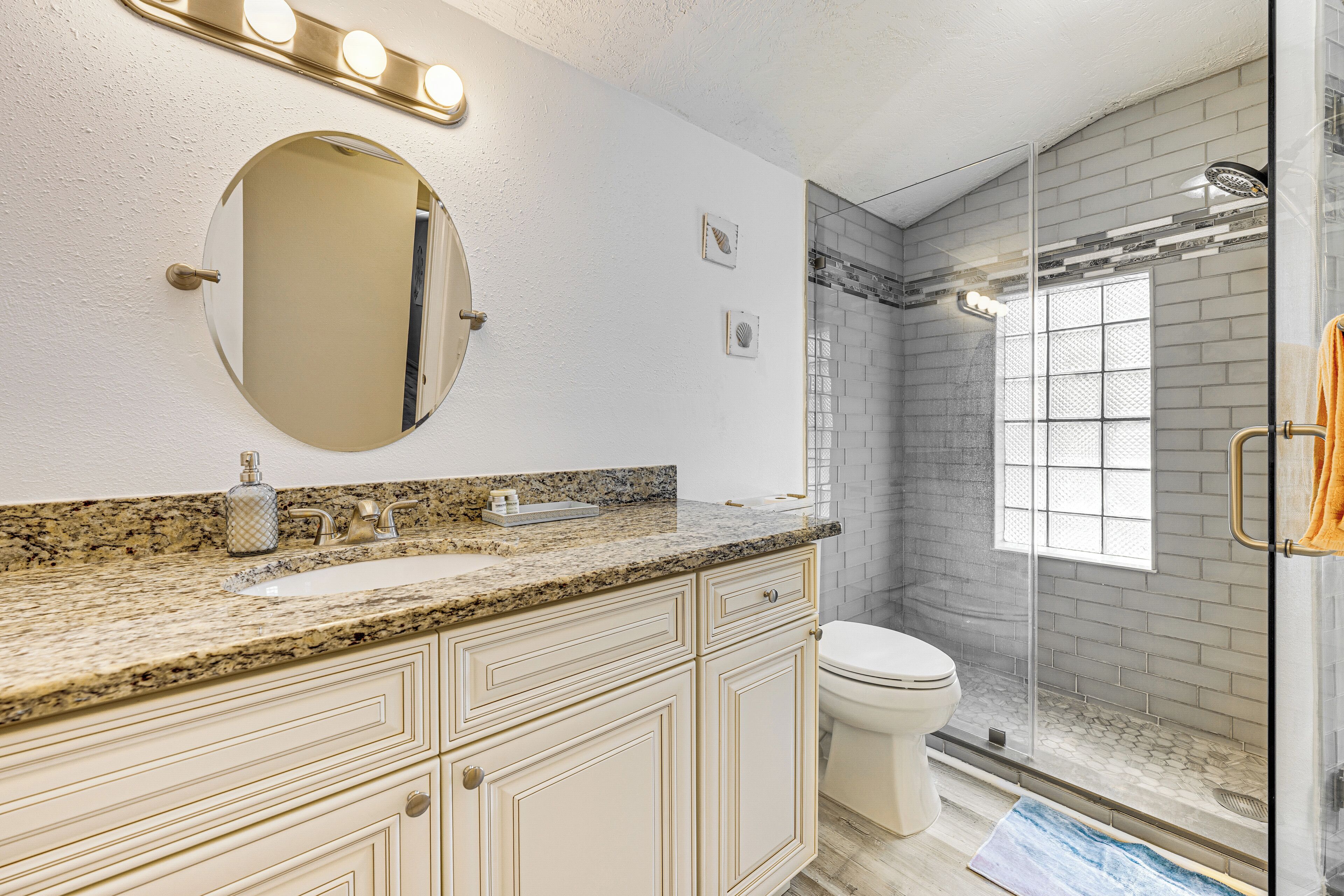 Un baño con encimera de granito, lavamanos, inodoro, ducha con puerta de cristal y espejo con luces arriba.