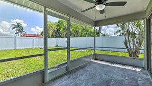A covered patio area with a ceiling fan, large windows, and a view of a grassy yard with a white fence and palm trees.