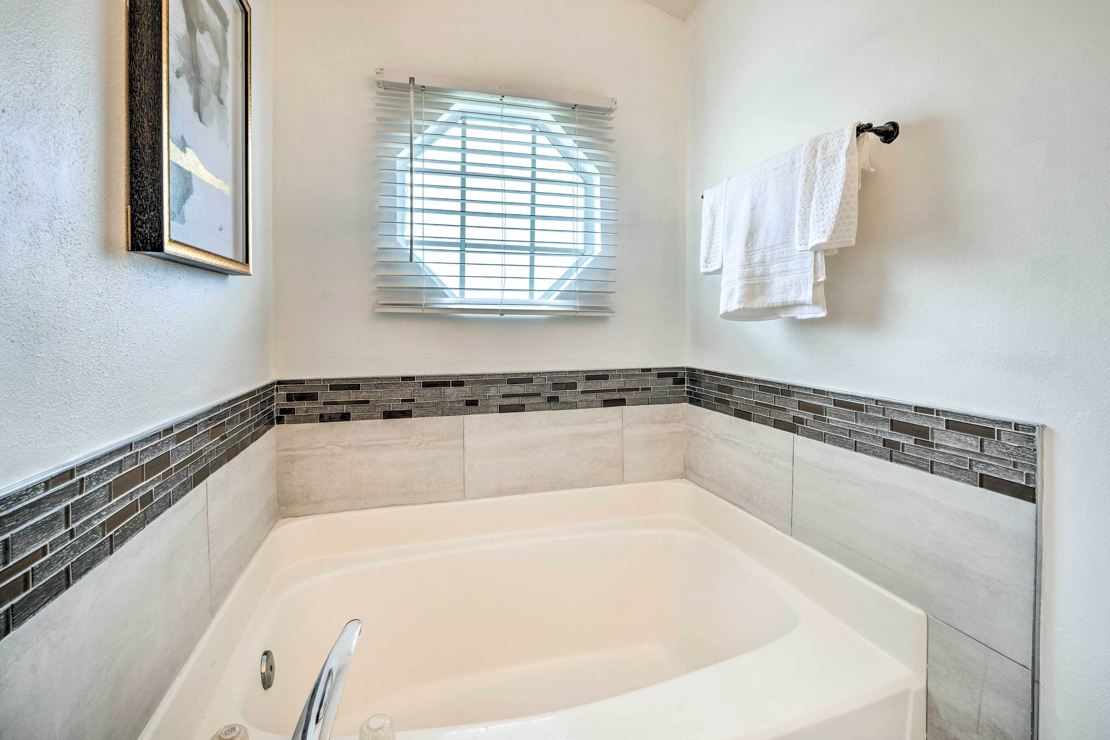 A bathroom with a large bathtub, a window with blinds, and a tiled wall with a brick pattern.