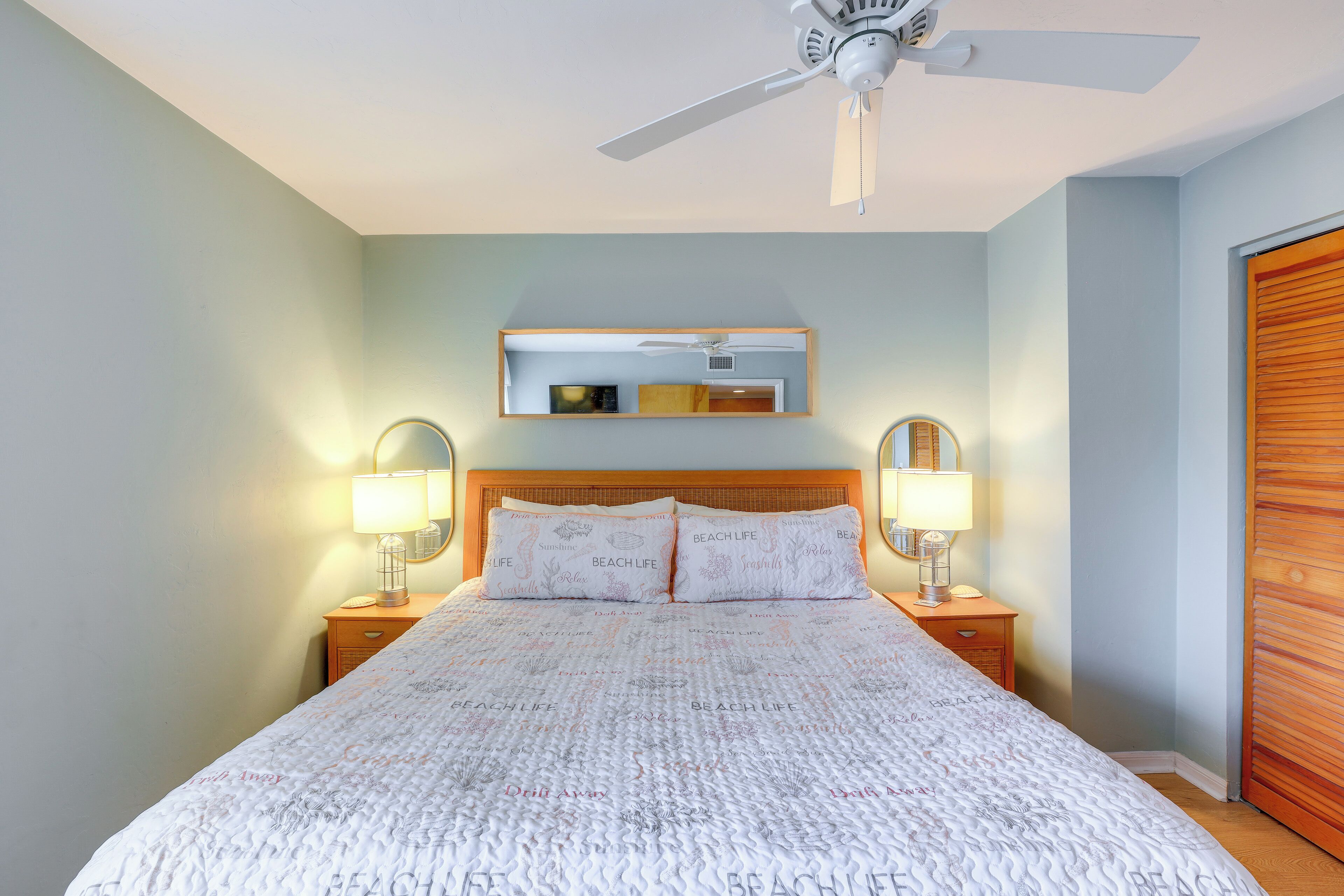 Un lit bien fait, recouvert d’une couvre-lit à motifs, deux lampes de chevet, un ventilateur de plafond, une tête de lit en bois, un miroir au-dessus du lit et une porte en bois sur le côté droit.