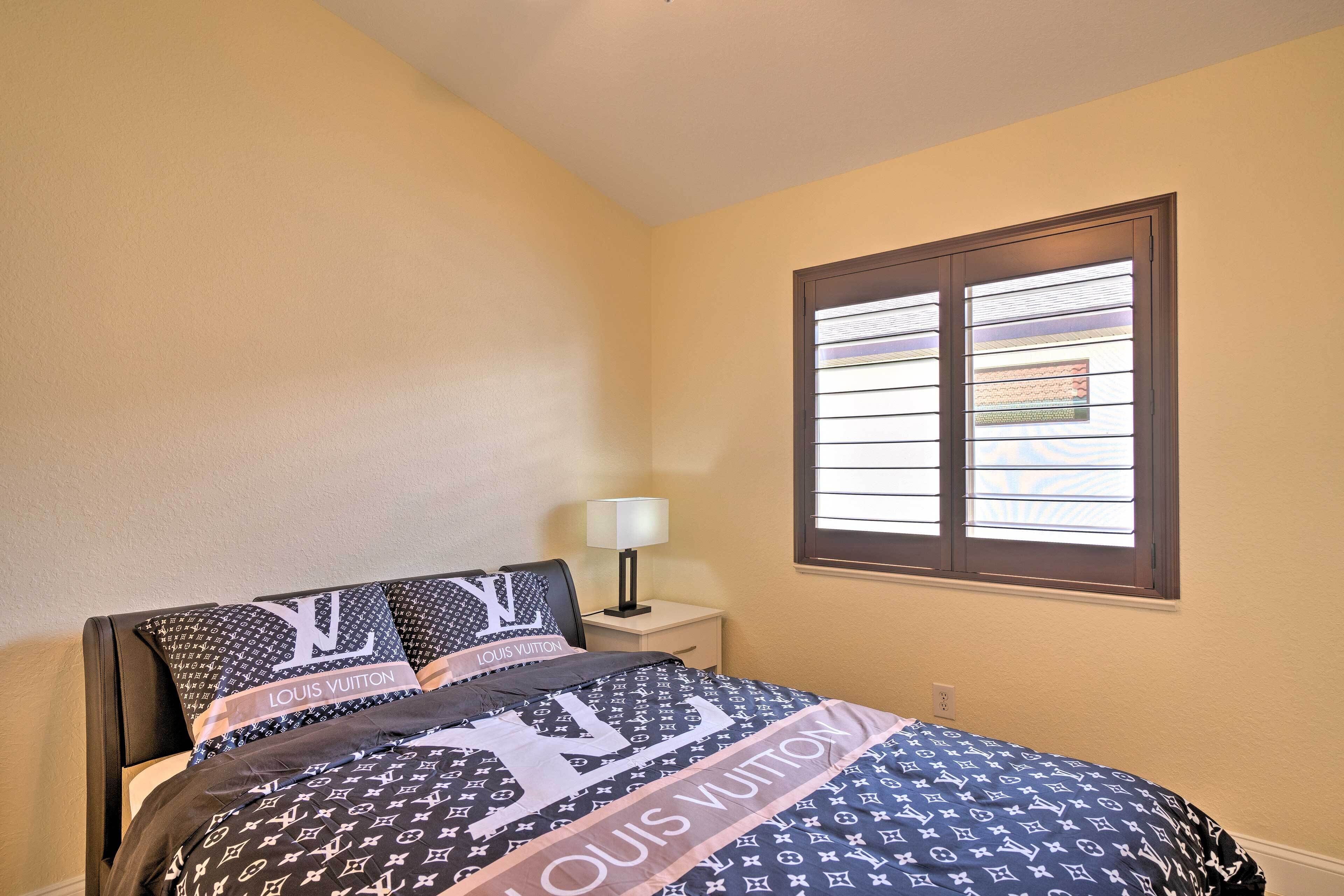 Un dormitorio con una cama con ropa de cama Louis Vuitton, una mesita de noche con una lámpara y una ventana con persianas.