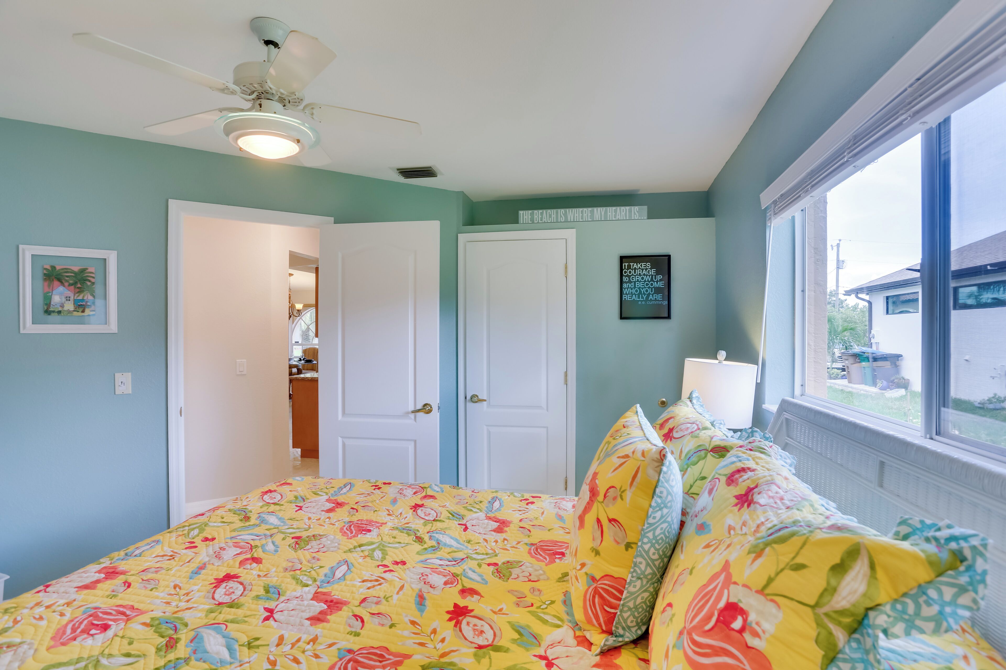 A bedroom with a floral bedspread, a ceiling fan, and a window with a view of the outside.
