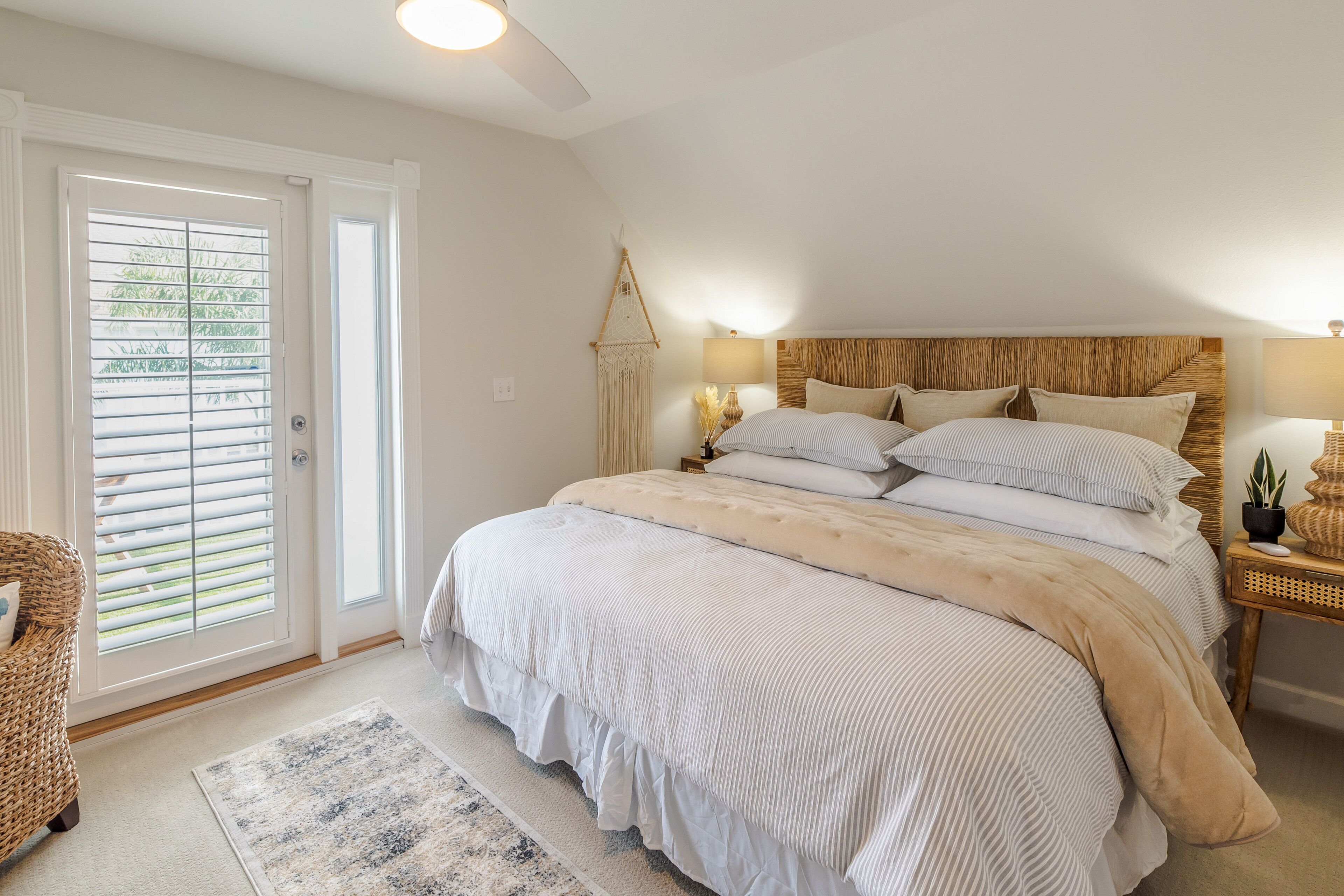 Een slaapkamer met een groot bed, een nachtkastje met lamp, een rieten stoel en een deur met zonwering.