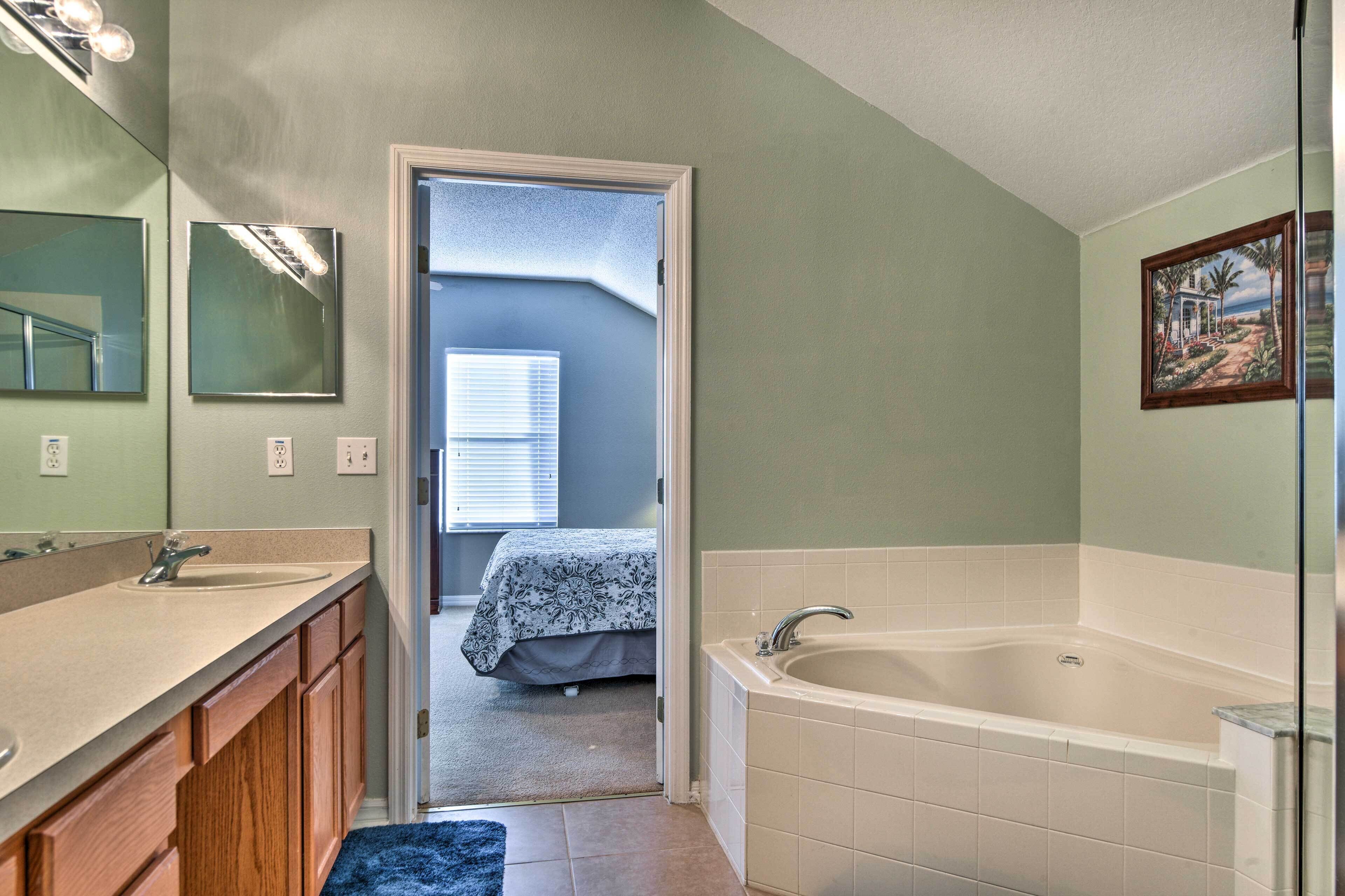 Un baño con lavamanos, espejo y bañera. Hay una puerta que da a un dormitorio con cama y ventana.