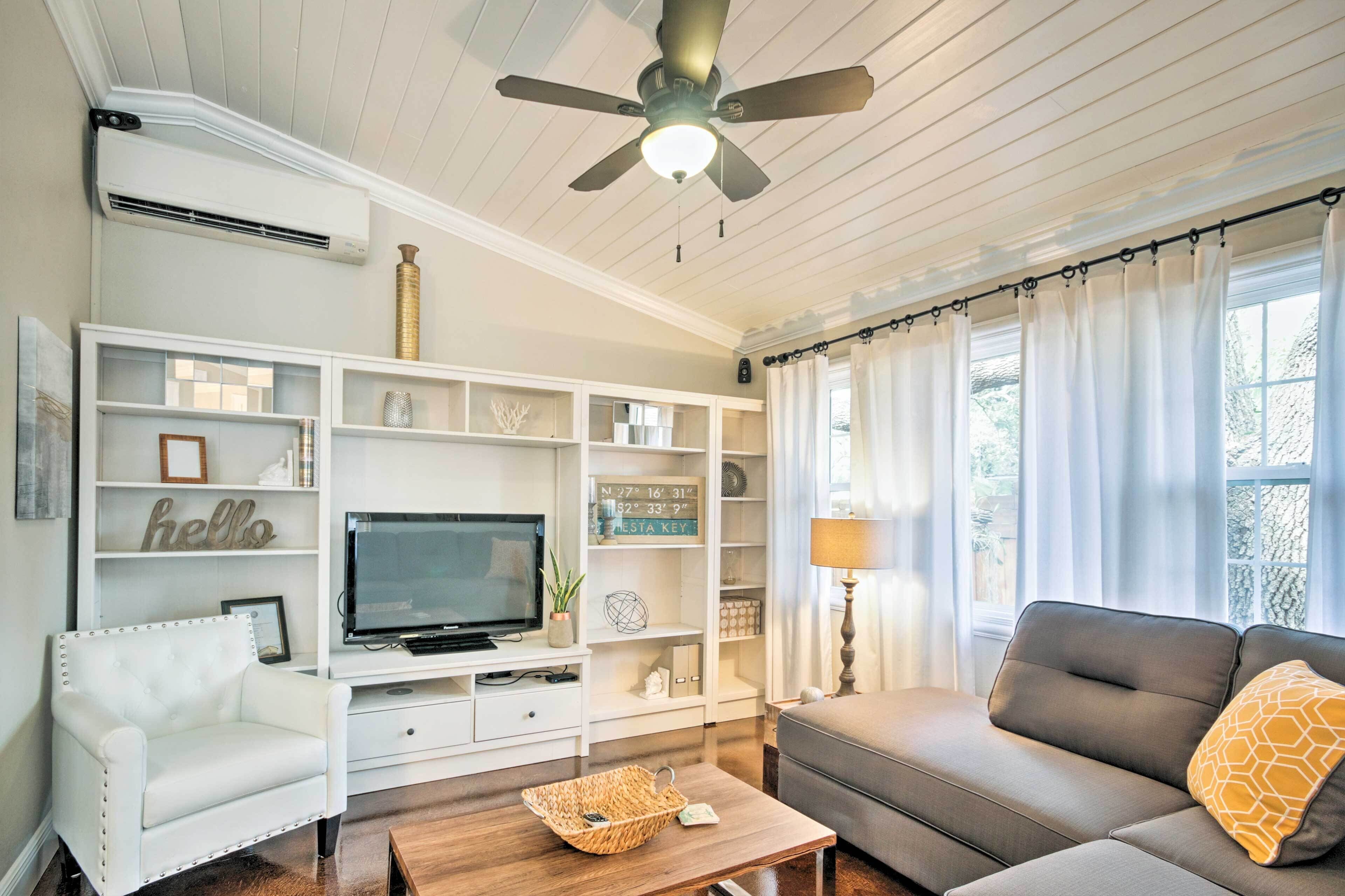 A living room with a white sofa, a grey sofa, a white armchair, a TV on a white entertainment unit, and a ceiling fan.