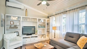 Una sala de estar con un sofá blanco, un sofá gris, un sillón blanco, un televisor sobre un mueble blanco y un ventilador de techo.