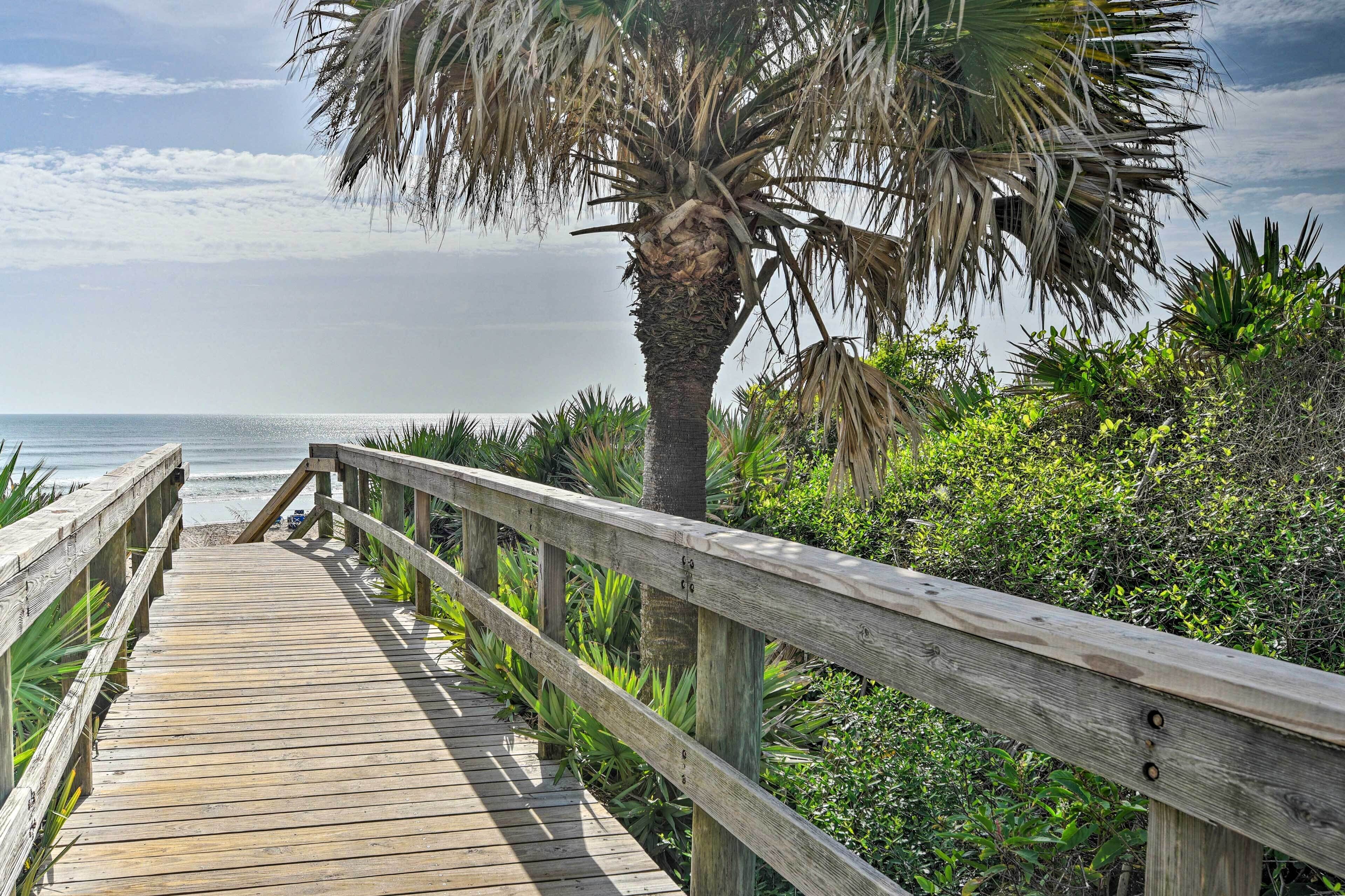 A wooden boardwalk leading to a beach with a palm tree and dense greenery.