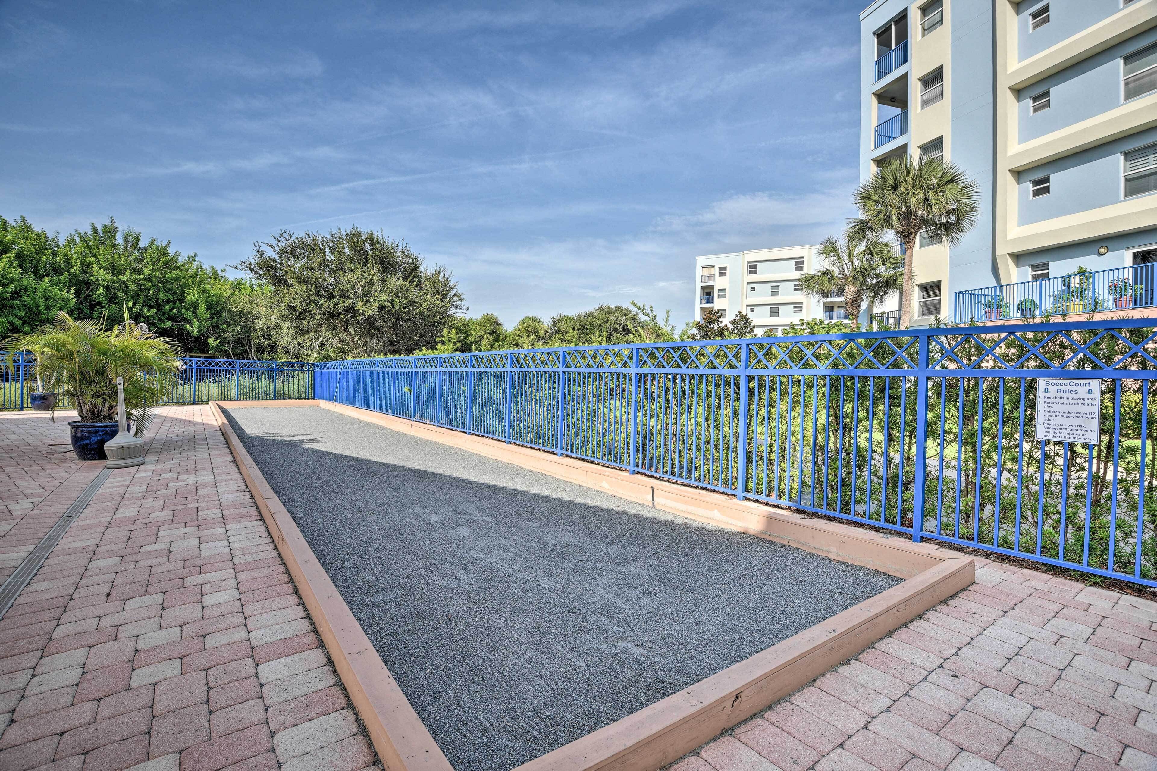 Un camino pavimentado con una cerca de metal azul, un pequeño jardín con plantas en macetas y un edificio de varios pisos al fondo.