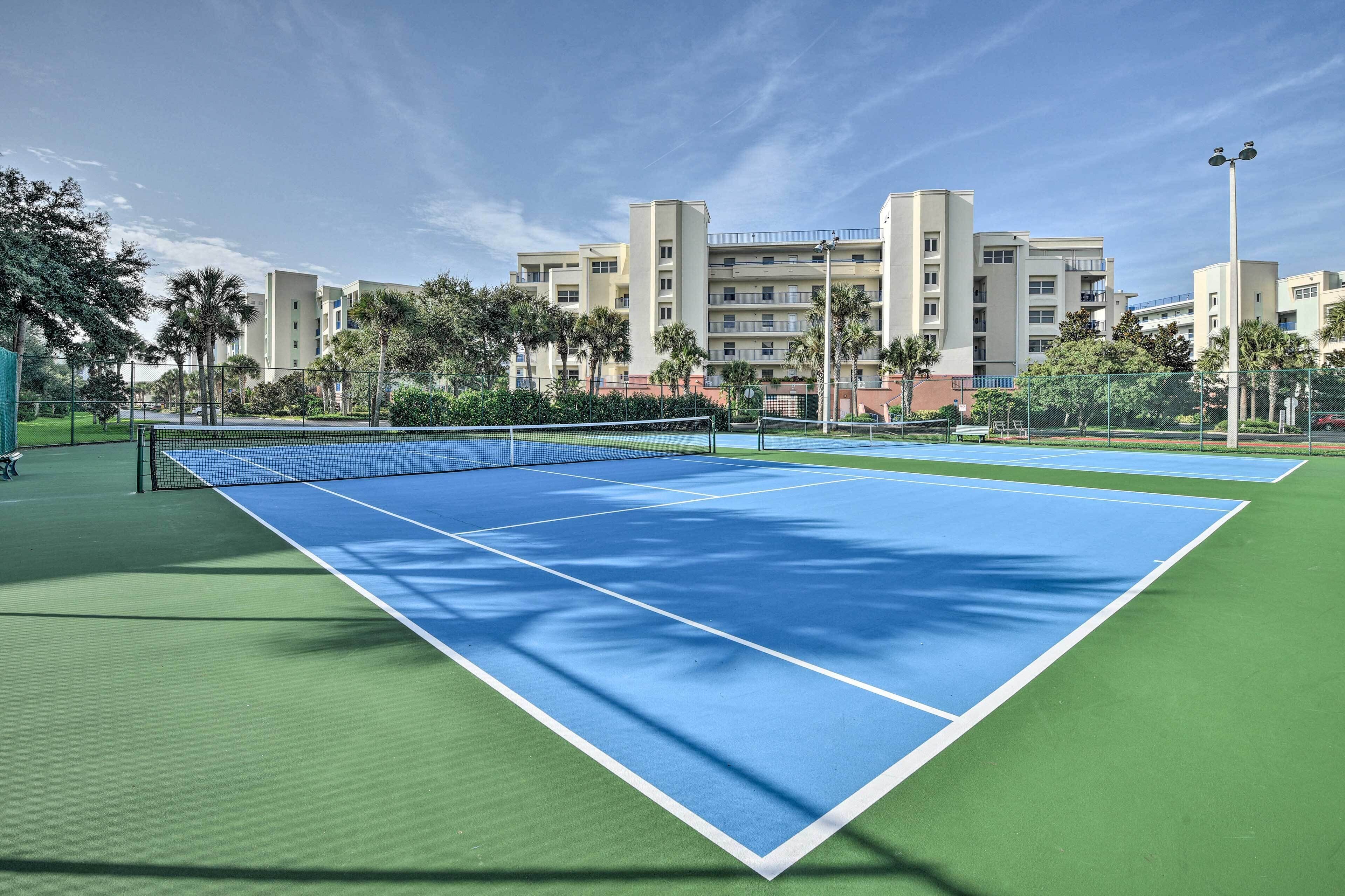 Ein Tennisplatz mit Netz, umgeben von Grünflächen und Gebäuden.