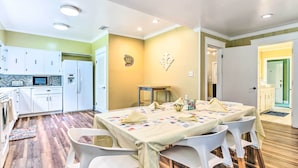 Uma sala de jantar com uma mesa posta para quatro pessoas, cadeiras brancas e uma cozinha ao fundo.
