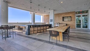 A spacious outdoor area with a long wooden table and multiple bar stools, a modern kitchen area, and a large window providing a view of the city skyline. - Chic Miami Condo w/ Pool, 6 Mi to South Beach (Miami)