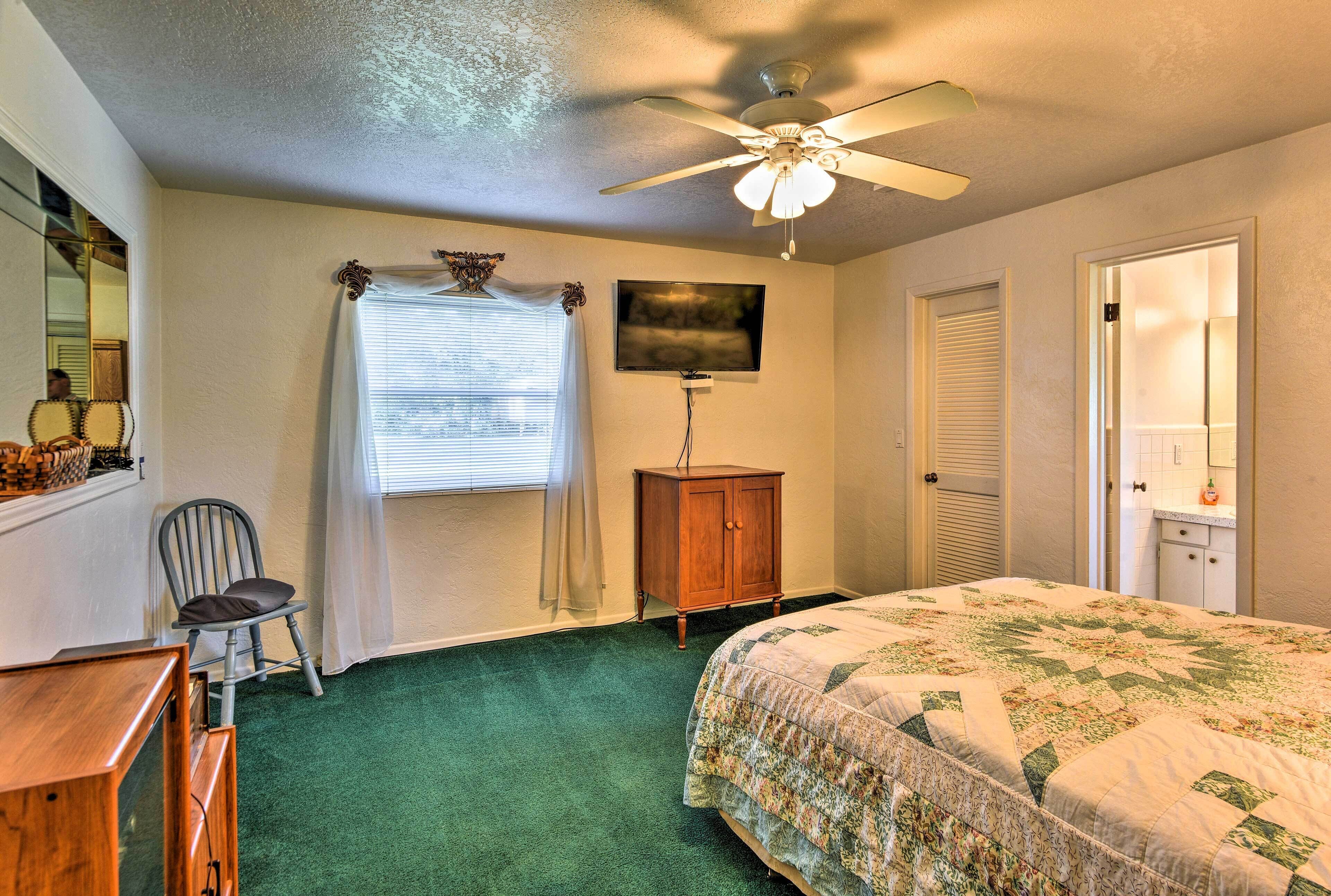 A bedroom with a green carpet, a bed with a floral quilt, a wooden nightstand, a chair, a ceiling fan, a TV, and a window with curtains.