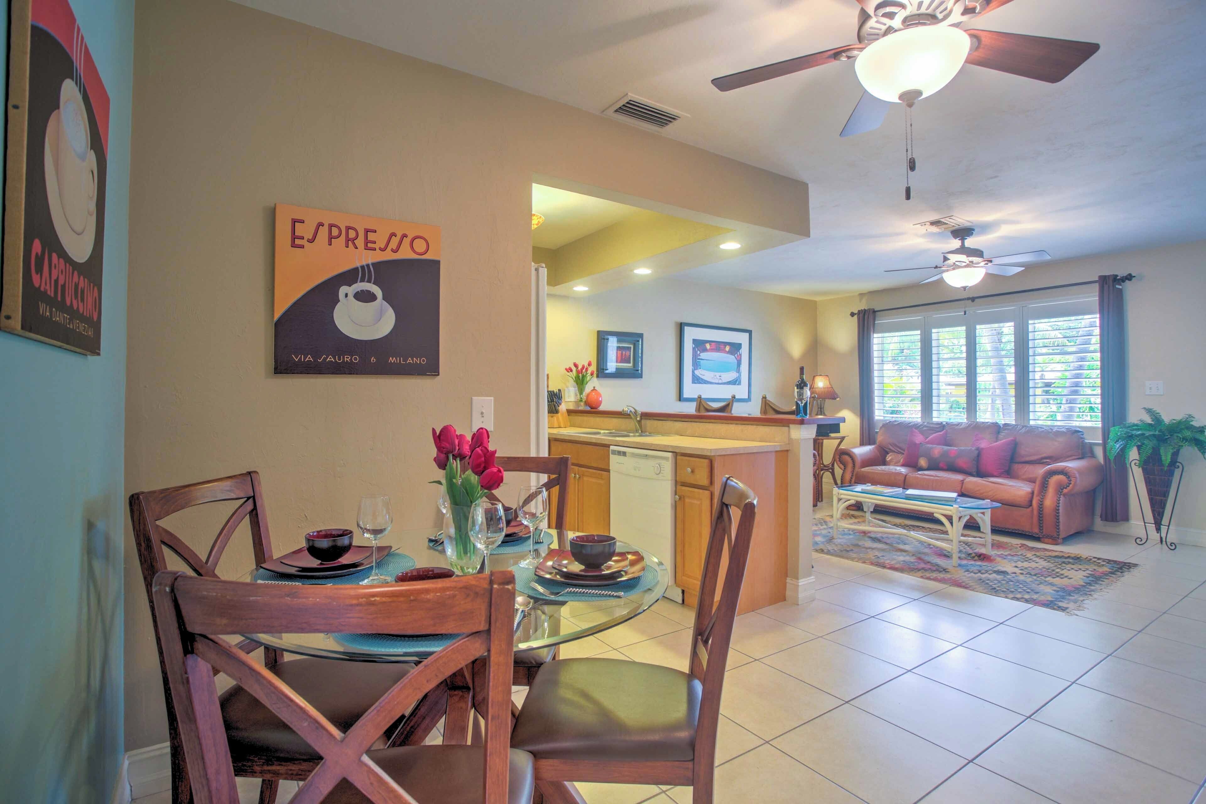 A dining area with a round glass table, wooden chairs, and a vase of red flowers. There is a kitchen in the background with a countertop and a refrigerator. A ceiling fan is visible above the dining area.