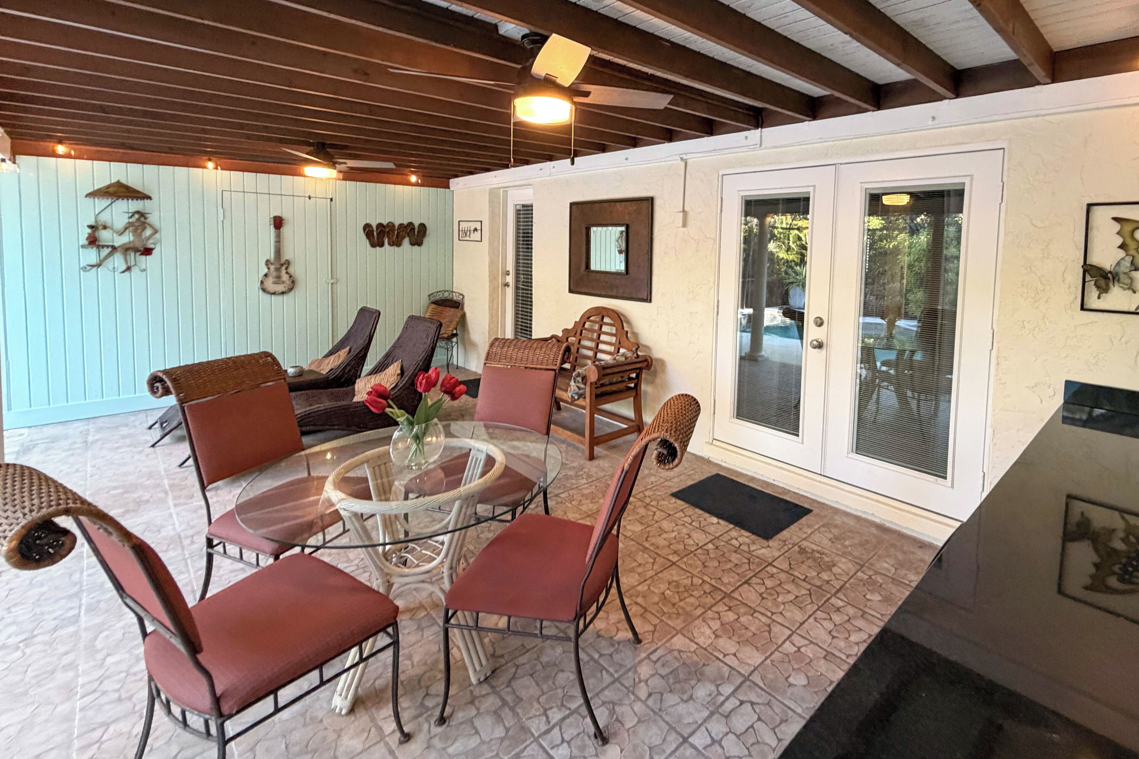 Un patio techado con una mesa de vidrio rodeada de sillas, un sofá y una pared decorativa con objetos colgados.