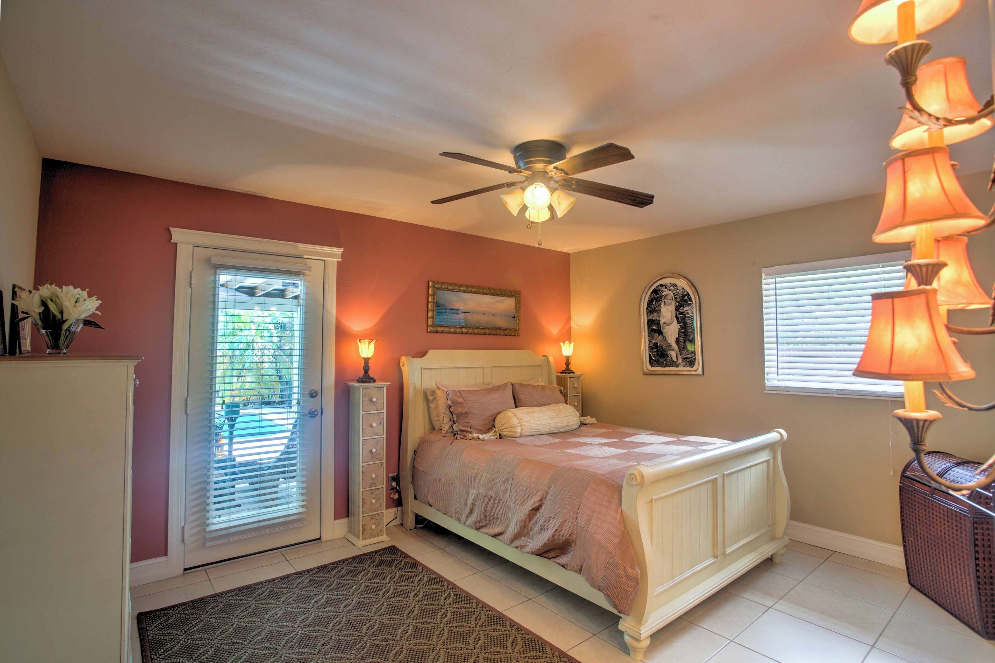 A bedroom with a bed, ceiling fan, bedside lamps, a window with blinds, and a door with a view.