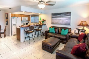 A modern living room with a leather sofa, a dining area with bar stools, a kitchen with a microwave, and a ceiling fan.
