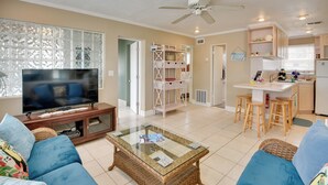 A living room with a television, blue sofas, a glass coffee table, and a kitchen area with stools and a refrigerator. - Cozy Ormond Beach Bungalow: Steps to the Sand (Ormond Beach)