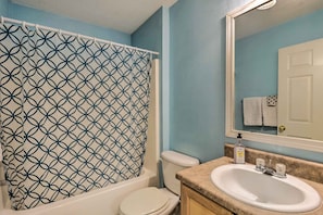 Ein Badezimmer mit gemustertem Duschvorhang, Toilette, Waschbecken mit Spiegel darüber und einer Handseifenflasche auf der Ablagefläche.