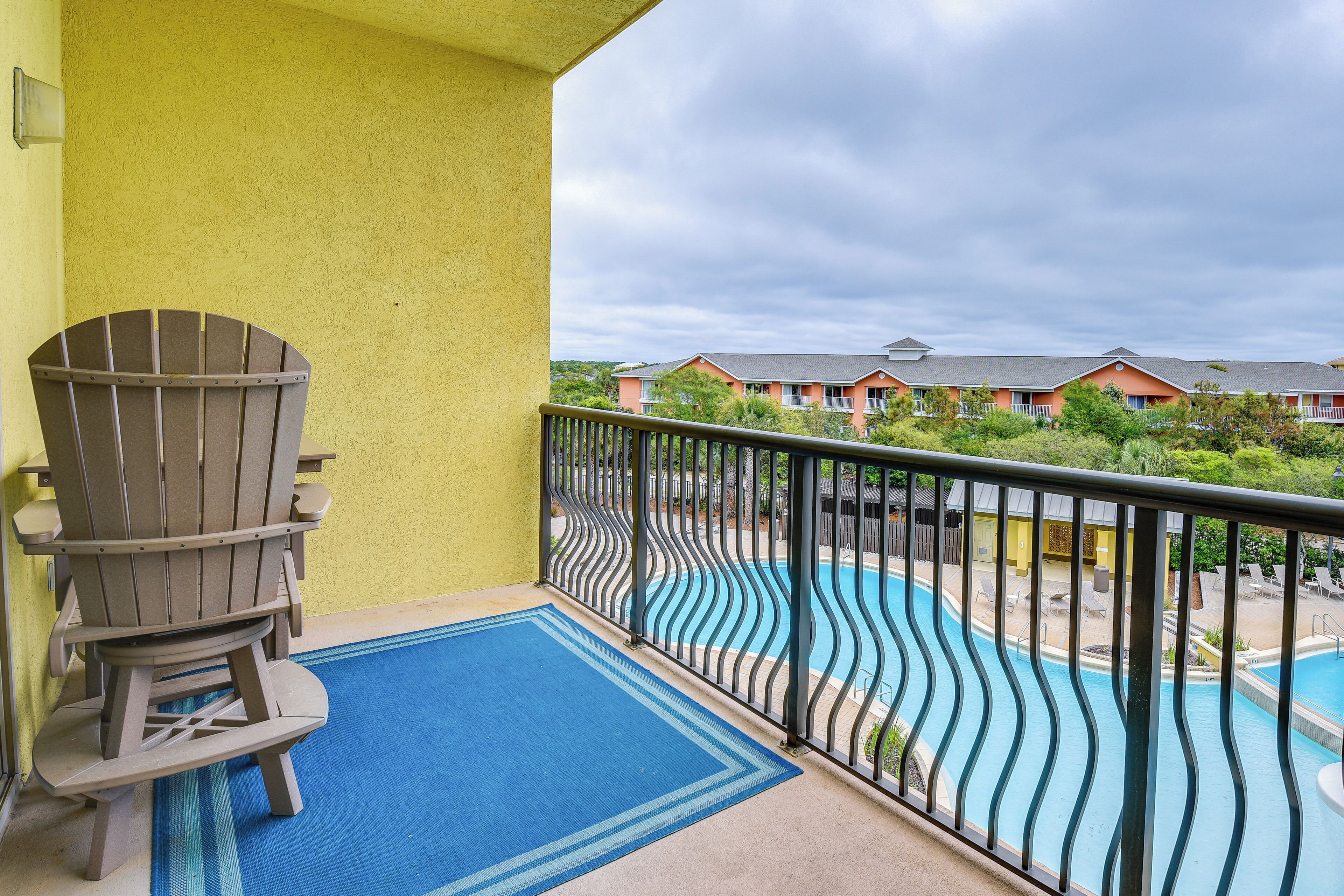 A balcony with a Adirondack chair, a blue mat, and a view of a swimming pool and buildings.