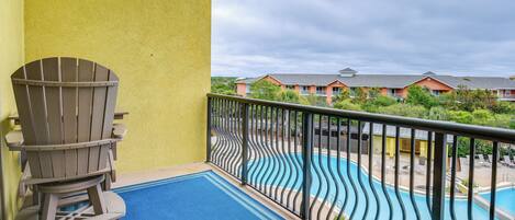 A balcony with a Adirondack chair, a blue mat, and a view of a swimming pool and buildings.
