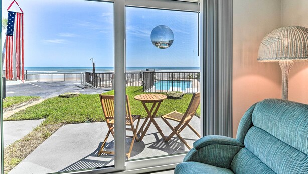A room with a large sliding glass door leading to a balcony with a view of the beach and pool. There is a round mirror on the door, a wicker lamp, and a blue sofa. - Daytona Beach Retreat w/ Views & Pool Access! (Daytona Beach)