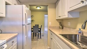 A kitchen with white cabinets, a stainless steel sink, and a dining area with chairs and a table. - Cozy Indian Rocks Beach Condo - Steps to Beach! (Indian Rocks Beach)