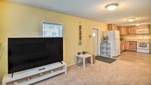 A living room with a flat-screen TV, a small white table, and a kitchen area with wooden cabinets and appliances. - Daytona Beach Home ~ 4 Mi to Daytona Speedway (Daytona Beach)