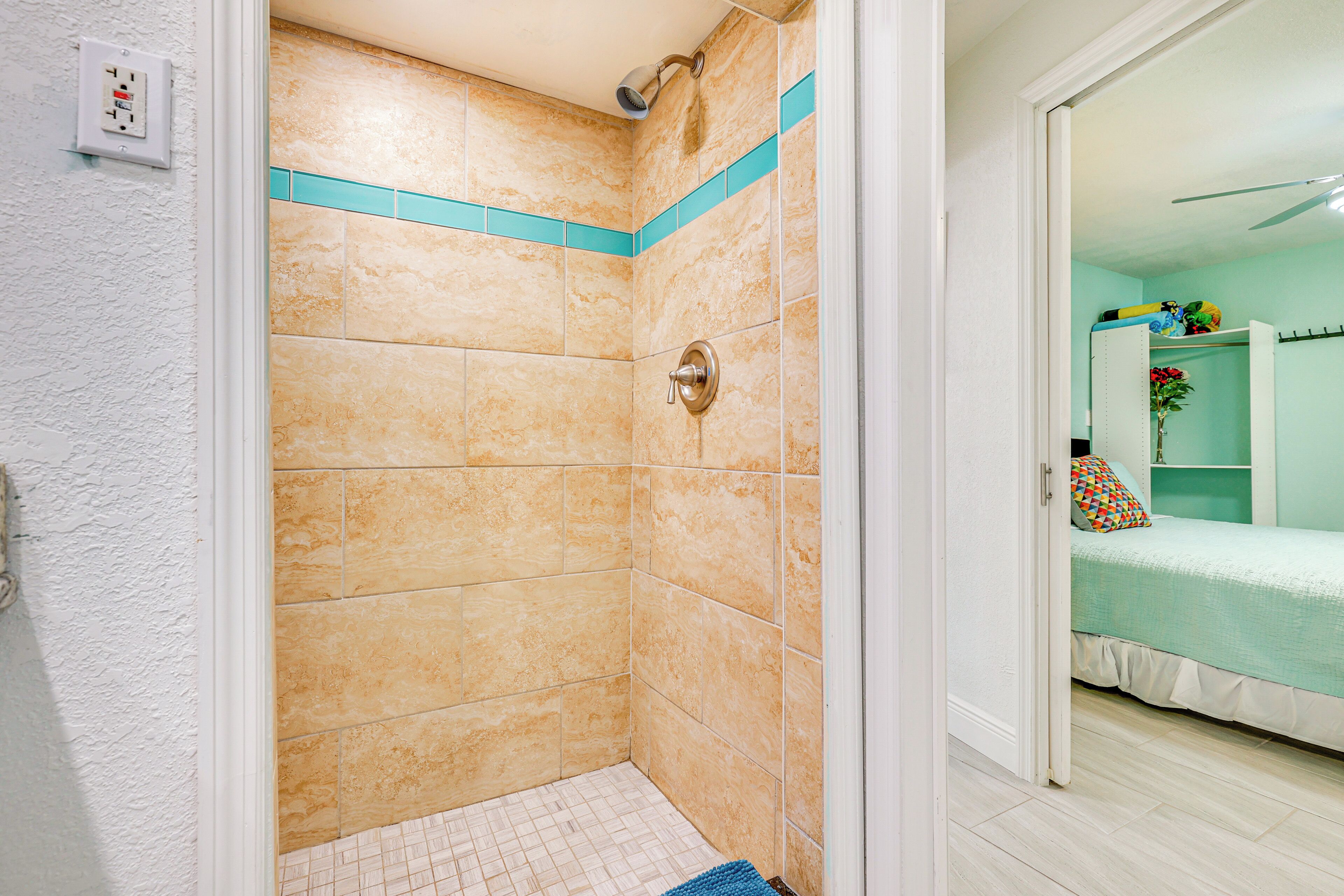 A bathroom with beige tiles, a shower, and a door leading to a bedroom with a bed and a shelf.