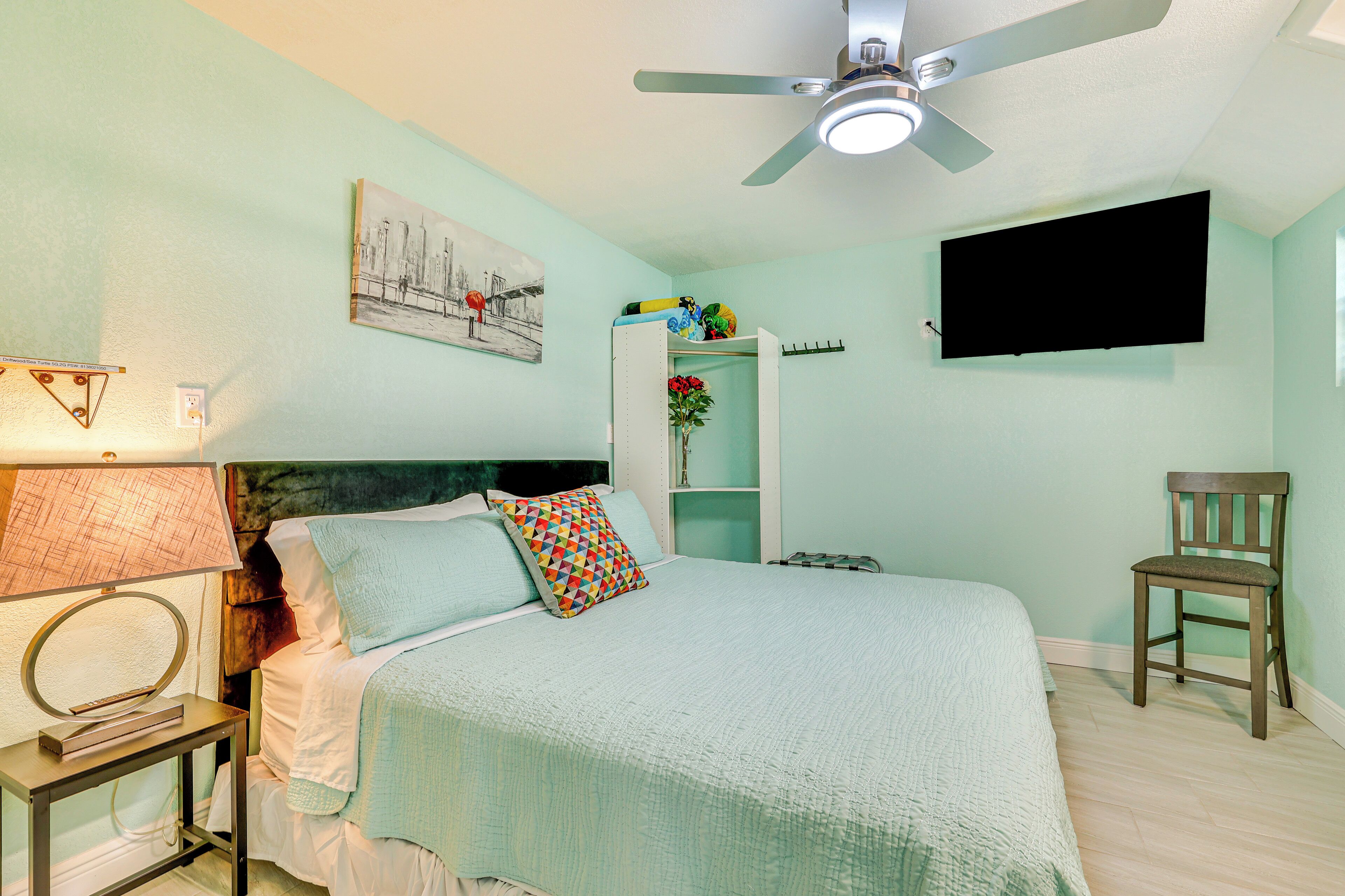 A bedroom with a bed, a chair, a TV, a ceiling fan, a lamp, a shelf with decorative items, and a painting on the wall.